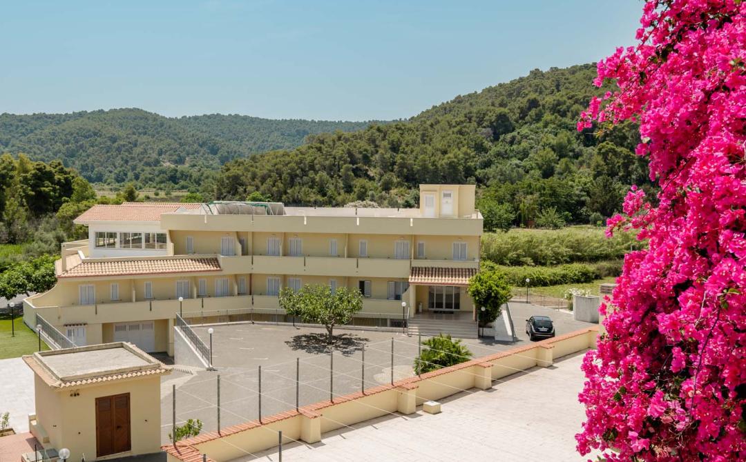 Edificio giallo tra colline verdi e fiori rosa vibranti, atmosfera serena e luminosa.