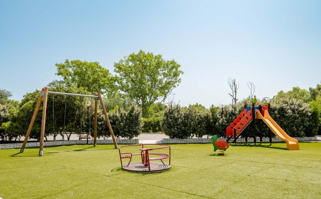 Parco giochi soleggiato con altalena, scivolo e giostra su prato verde.