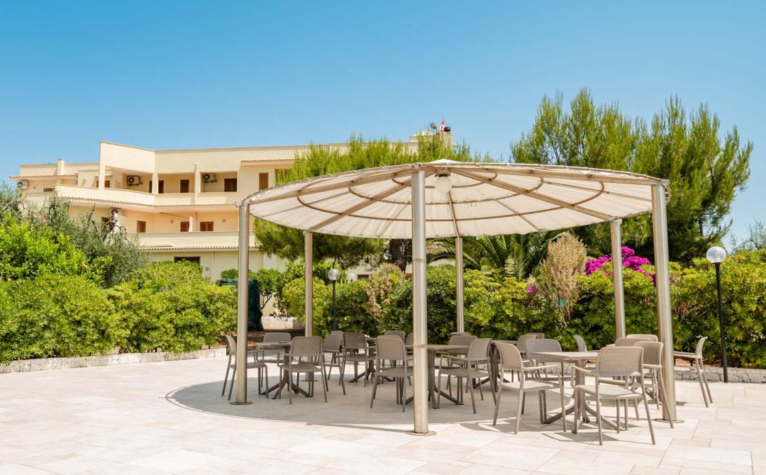 Gazebo soleggiato con sedie, circondato da verde lussureggiante e cielo azzurro.