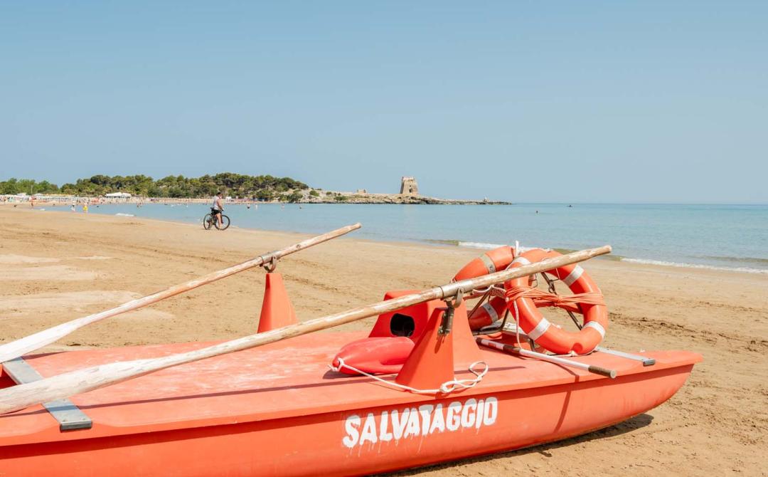 Spiaggia dorata, mare calmo, barca di salvataggio rossa, torre antica in lontananza.
