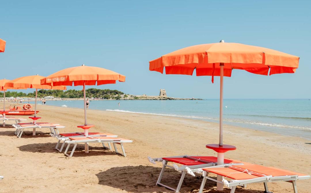 Spiaggia soleggiata con ombrelloni arancioni, mare calmo e torre in lontananza.